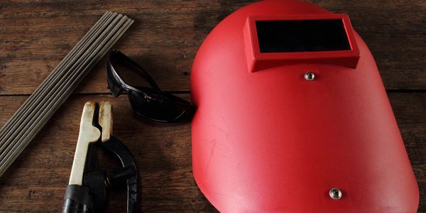 Welding gear including a red helmet, protective glasses, and welding rods on a wooden surface.