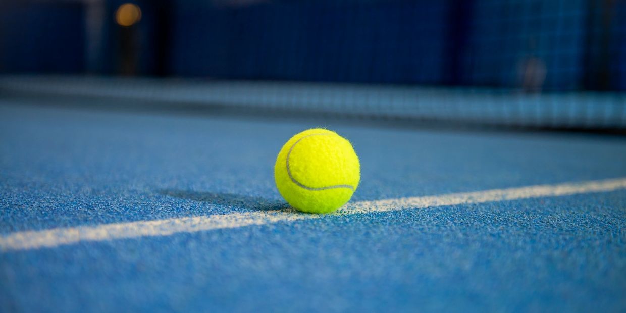 A bright yellow tennis ball sits on a blue tennis court near the white line.