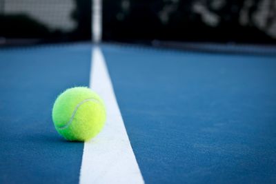 A tennis ball resting on the white line of a blue tennis court.