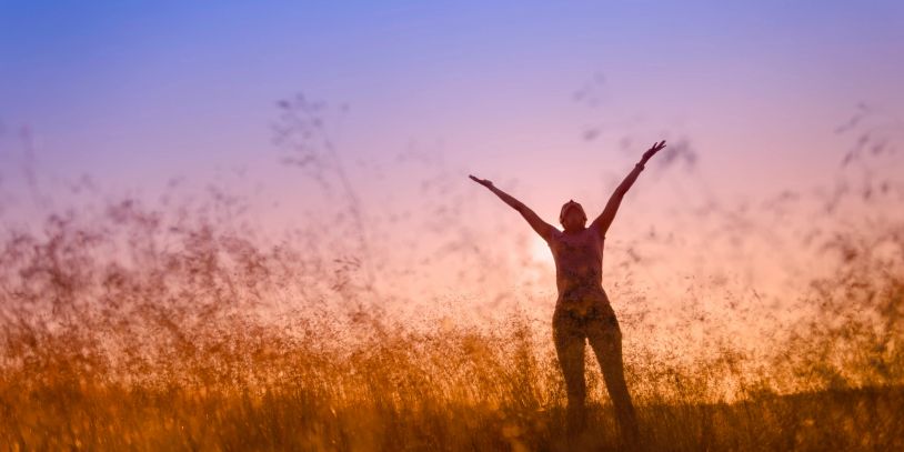 Person joyfully stands in a field at sunset with arms raised.