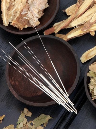 Acupuncture needles and dried herbs on dark wooden bowls and surface.