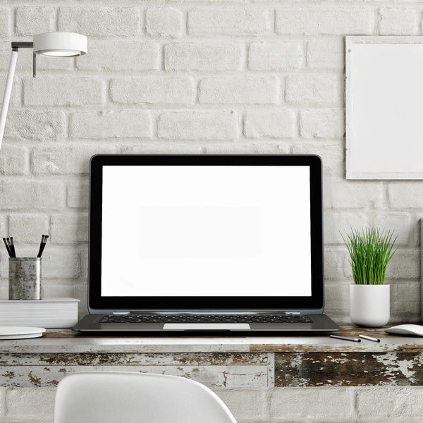Open laptop on desk with white brick wall background, a plant to the right and pens in a cup on left