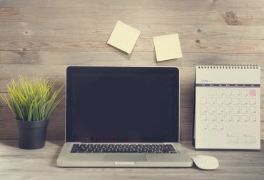 Workspace with laptop, calendar, plant, and sticky notes on wooden desk.