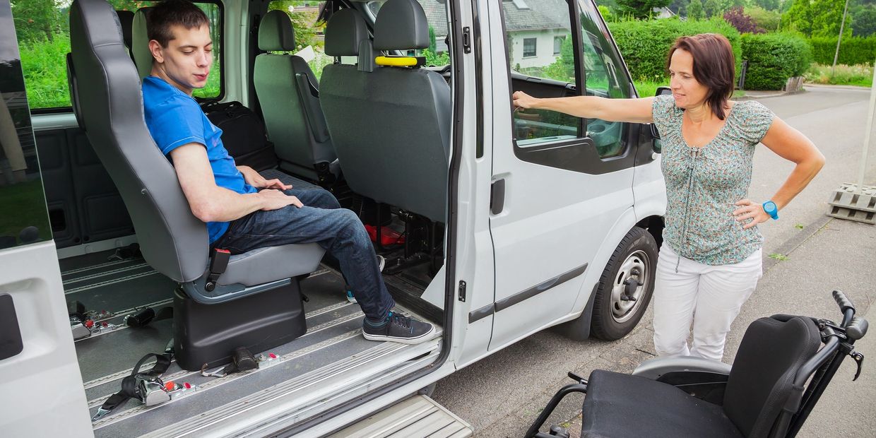 Man with special needs sitting inside a van beside a wheelchair and a caregiver