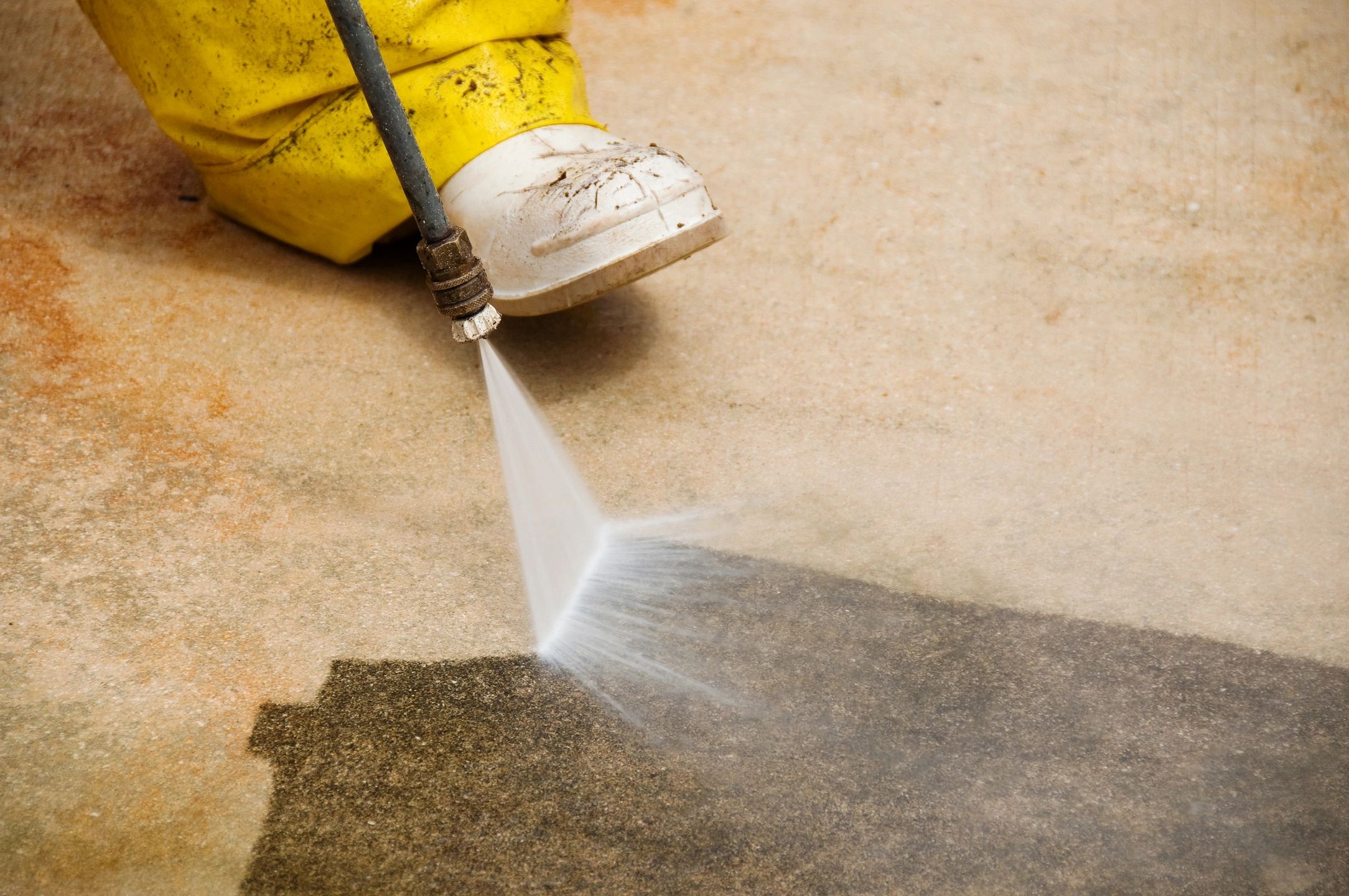 Person in yellow boots power washing a dirty concrete surface.
