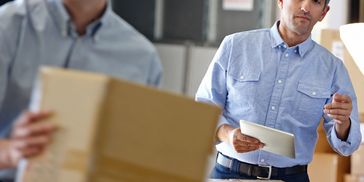 A man using a tablet while another carries a box in a warehouse.