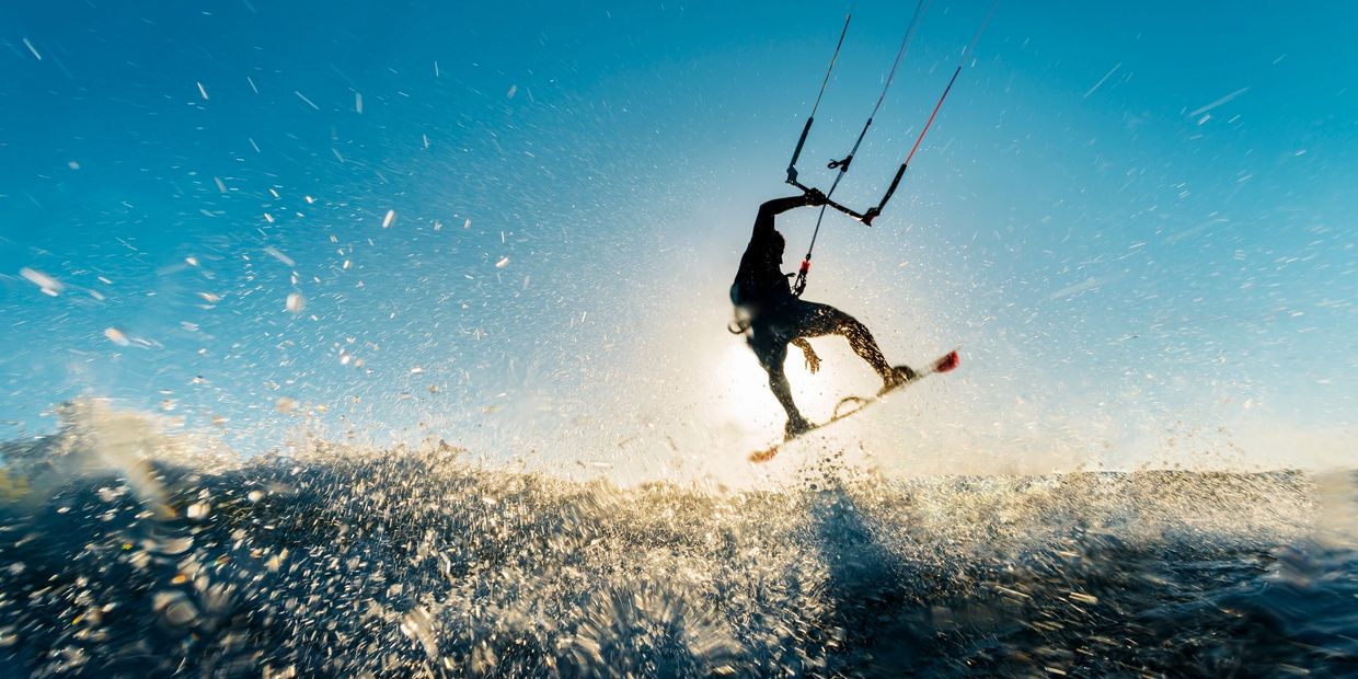 Silhouette of a kite surfer jumping over splashing waves against a bright sun.