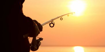 Person fishing with a rod at sunset over calm water.
