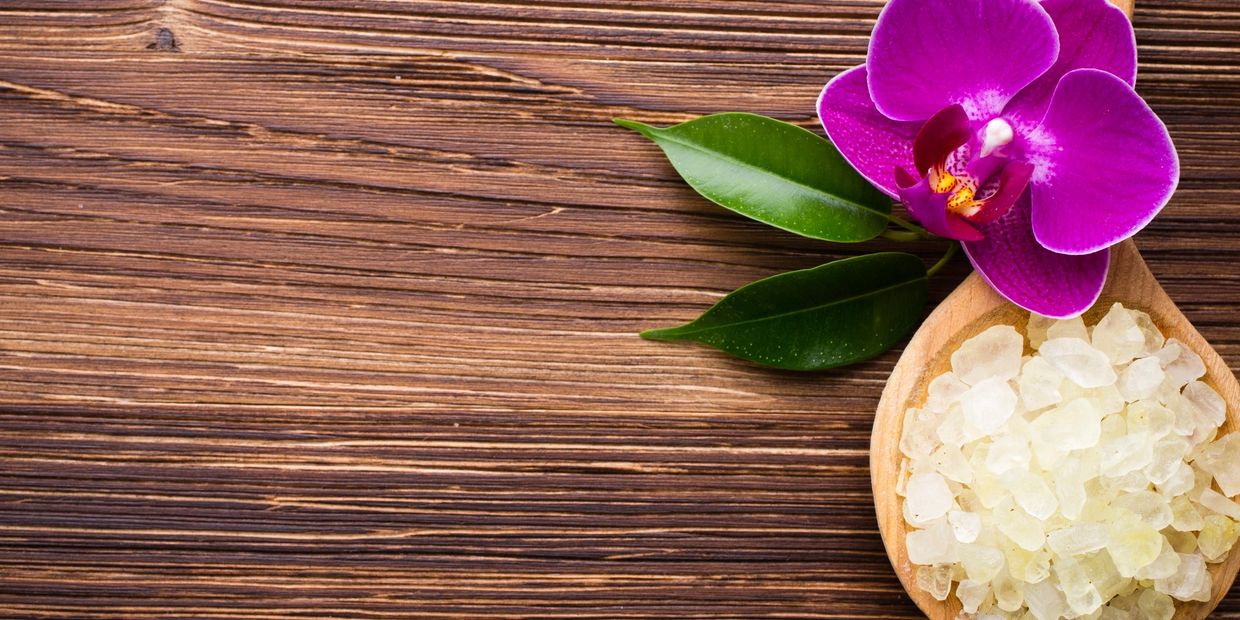 Wooden spoon with bath salts and a vibrant purple orchid on a wooden surface.