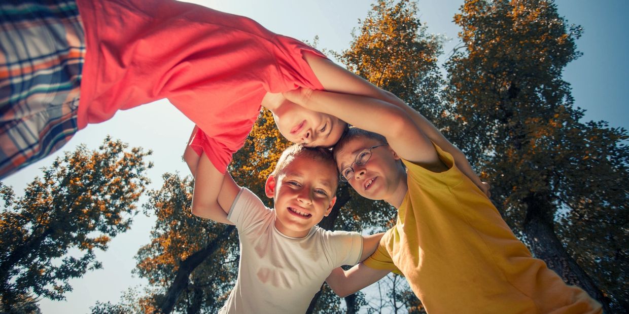 Three children smiling and embracing each other outdoors under tall trees.