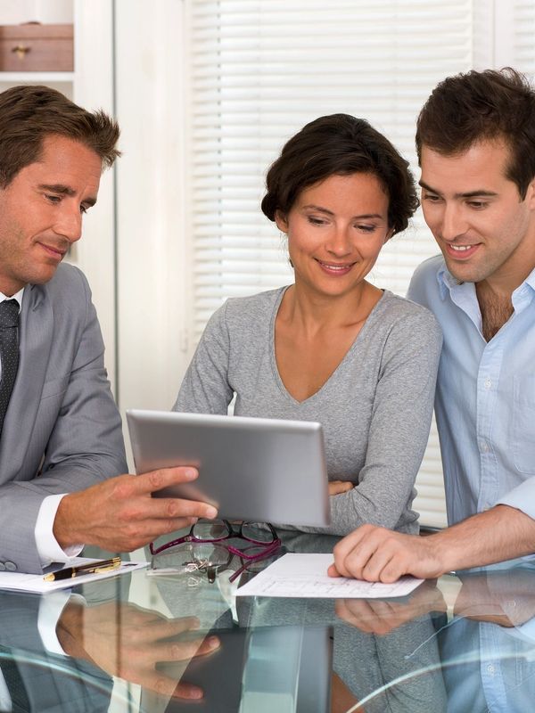 A man in a suit shows a tablet to a couple at a glass table.