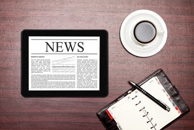 An open tablet lying on a table it says, "NEWS" There is an open notebook and a coffee mug.