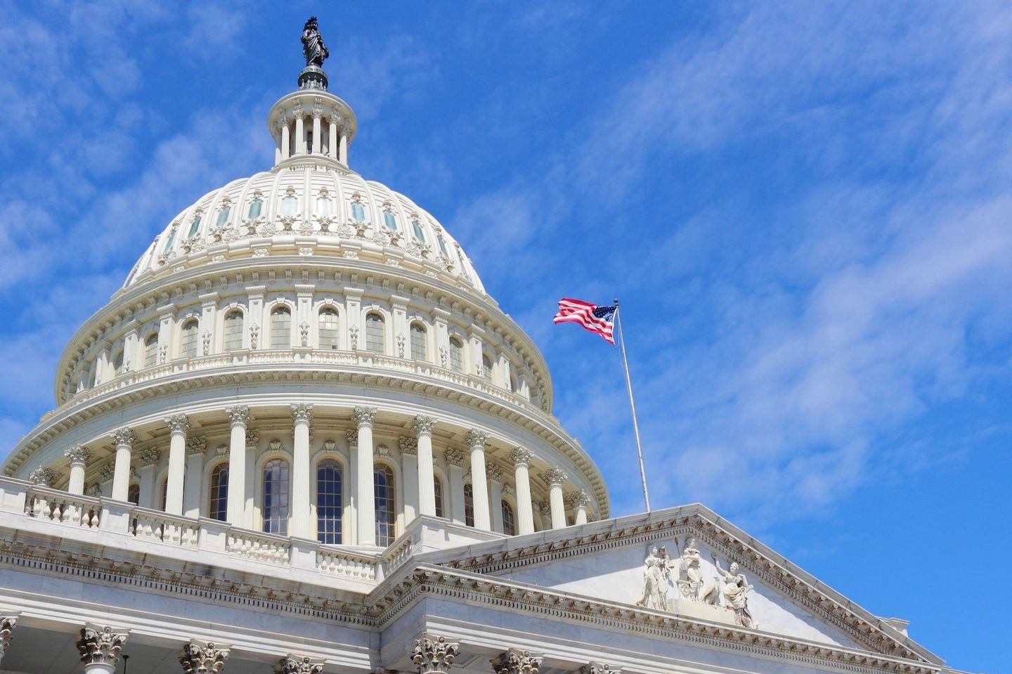 The United States Capitol dome with a waving American flag against a clear blue sky.