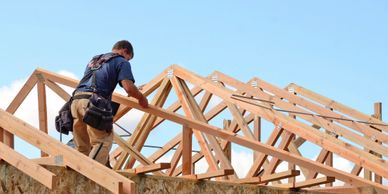 A worker constructing a wooden roof frame under clear blue sky.