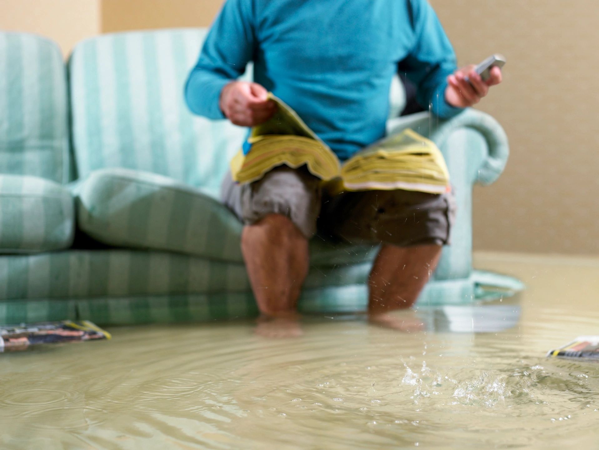 A person sitting on a couch with a book and phone in hand while the floor is flooded