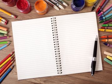 Open notebook with blank pages surrounded by colored pencils, paint, and a pen on a wooden table.