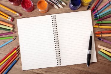 Open notebook surrounded by colorful art supplies on a wooden surface.