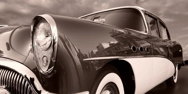 Sepia-toned close-up of a vintage classic car highlighting its headlight and curved fender.