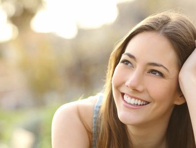 A smiling woman with long brown hair outdoors in soft natural light.