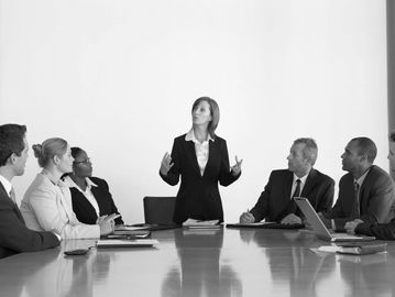 A business meeting with suited people listening to a person speaking who is standing up