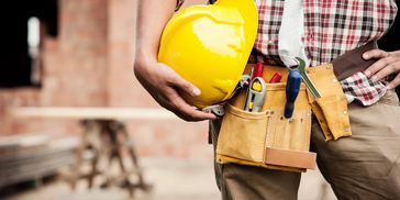 Construction worker holding a yellow helmet with tools in a belt.