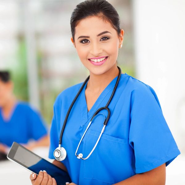 Smiling nurse in blue scrubs holding a tablet with a stethoscope around her neck.