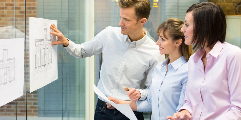 Three people reviewing architectural plans on a glass wall.
