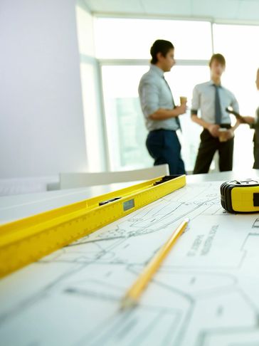 Yellow ruler and pencil on architectural plans with people talking.