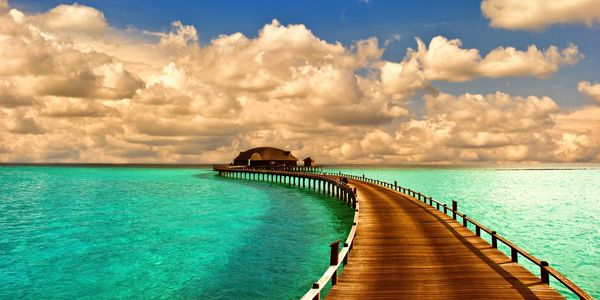 Curved wooden pier extending over turquoise ocean under cloudy sky.