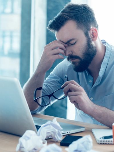 Stressed man taking off glasses while working on laptop.