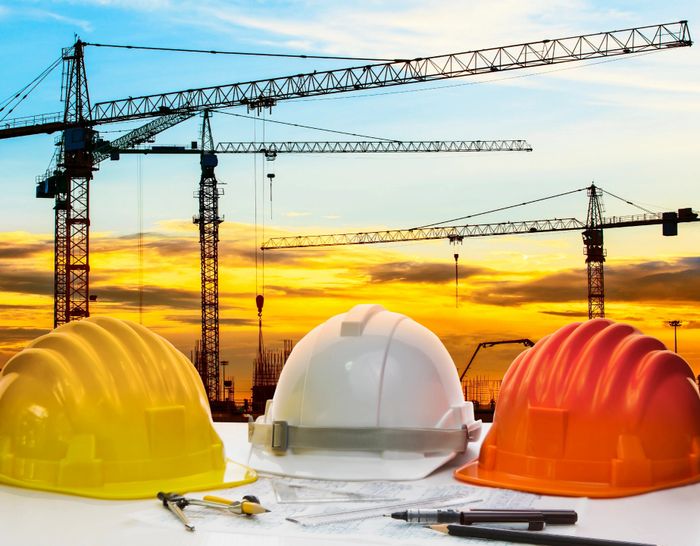 Three construction helmets and tools with cranes at sunset in the background.
