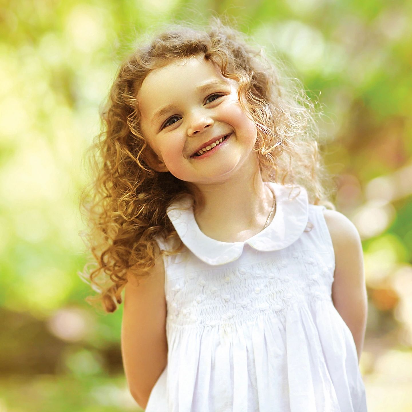 Smiling young girl with curly hair in a white dress outdoors.