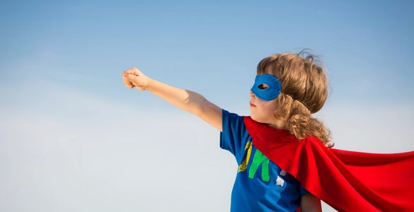 A child with curly hair in a blue shirt, eye mask, and red cape with arm extended to the sky