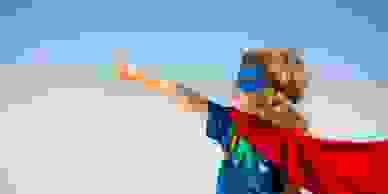 Little girl in superman costume pretending to fly, in front of a blue background.