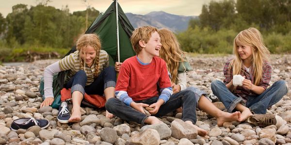 Four children sitting on rocky ground near a small green tent, laughing and enjoying each other's company.