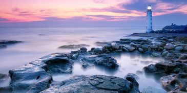 A lighthouse at sunset with rocky shore and calm sea.