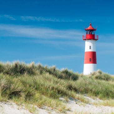 A red and white lighthouse stands tall against a clear blue sky by grassy sand dunes.