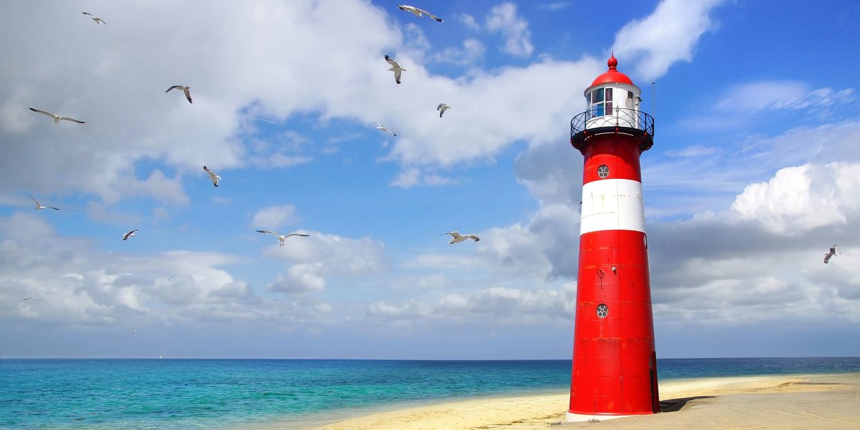 Red and white lighthouse on a sandy beach with seagulls flying above.