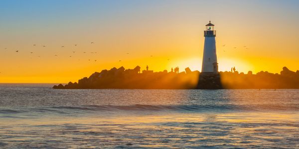 Lighthouse silhouette at sunset with people on rocky shore.