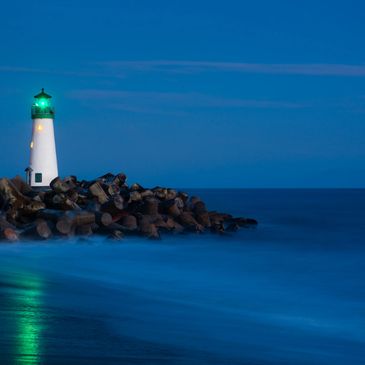 A lighthouse with a green light on a rocky shore at night.