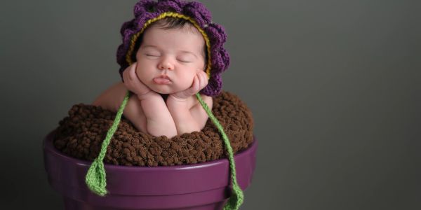 Sleeping baby wearing a purple knitted hat inside a purple flower pot.