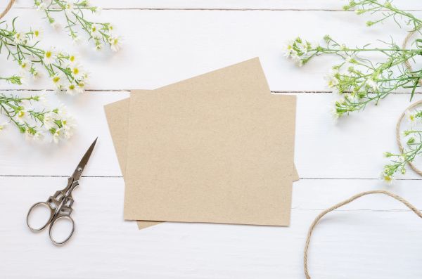 Blank brown cards with scissors and flowers on white wooden table.