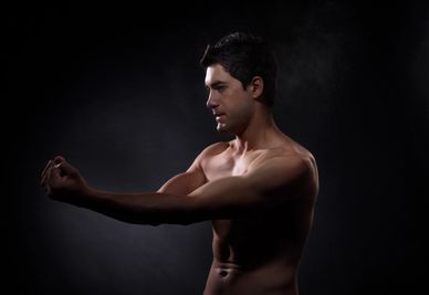 A shirtless man stretching his arm against a dark background.