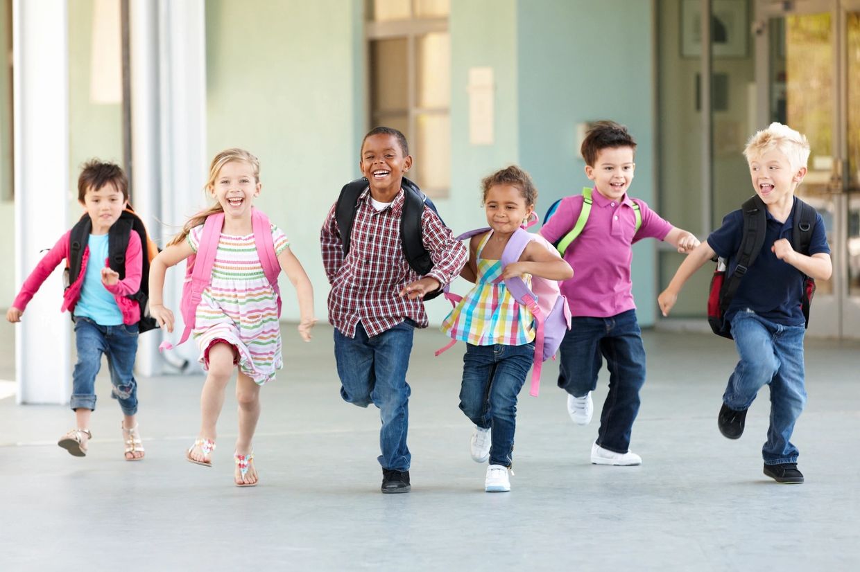 Elementary school students running together outside a school building while wearing backpacks
