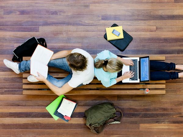 Two students studying together on a bench, one reading a book and the other using a laptop.