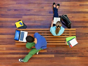 Two students sitting on a bench with their books and laptops out.