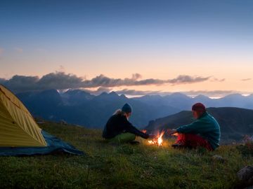 Two people sitting by a campfire near a tent in the mountains at sunset.