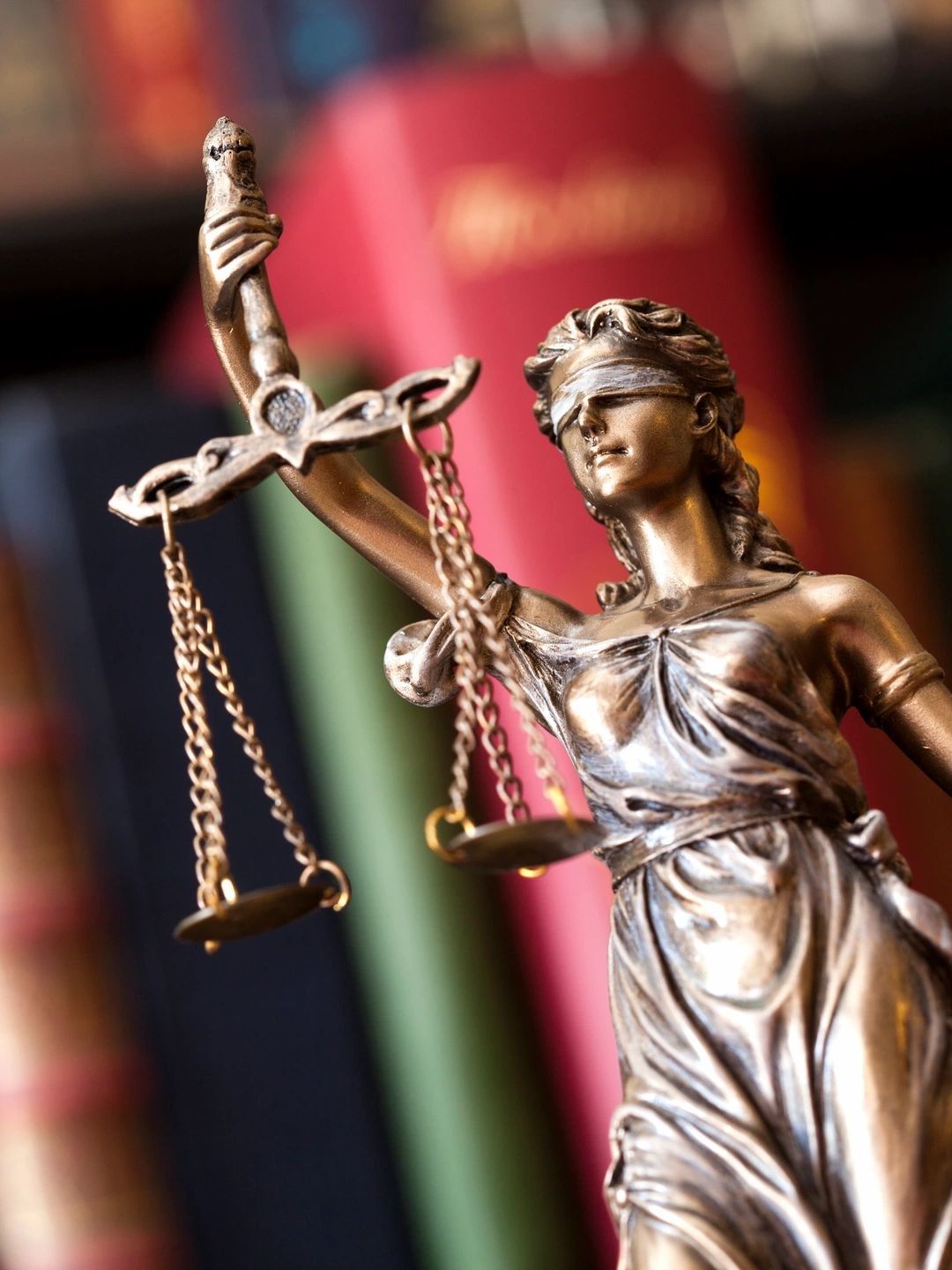 Close-up of Lady Justice statue holding scales with blurred books in background.