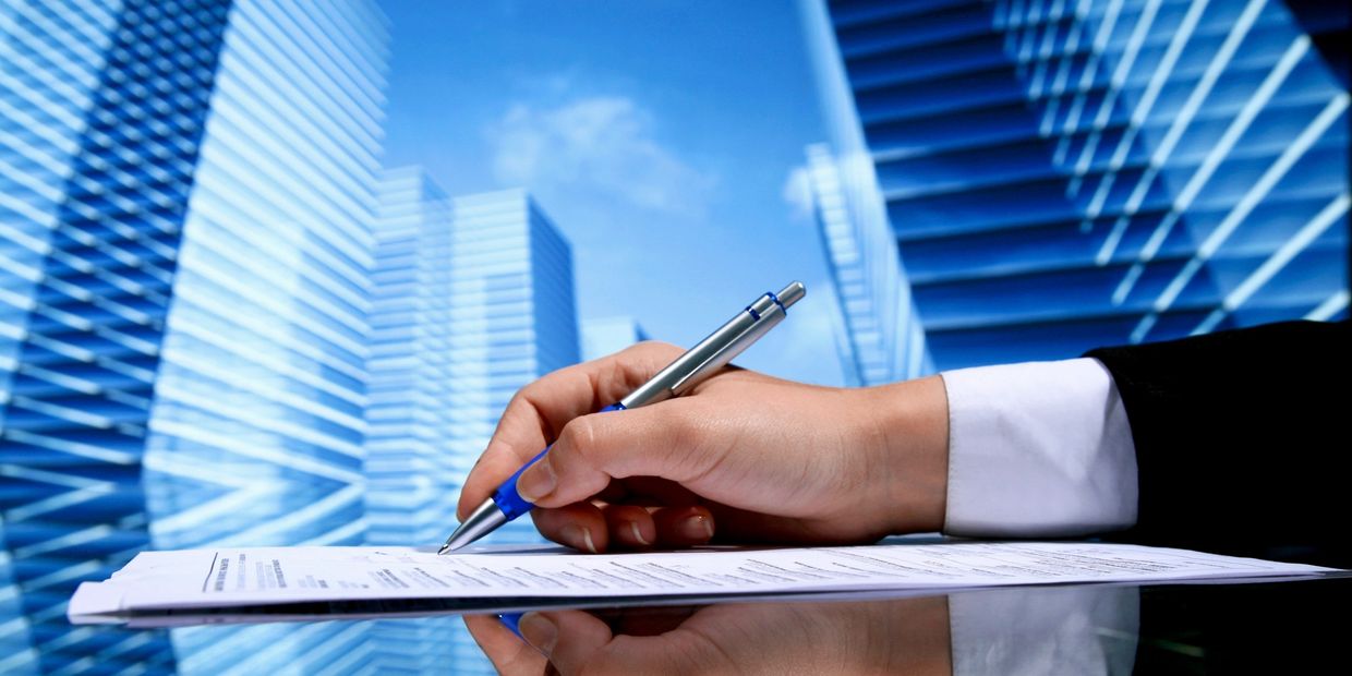 A hand in a business suit signing documents against a cityscape background.