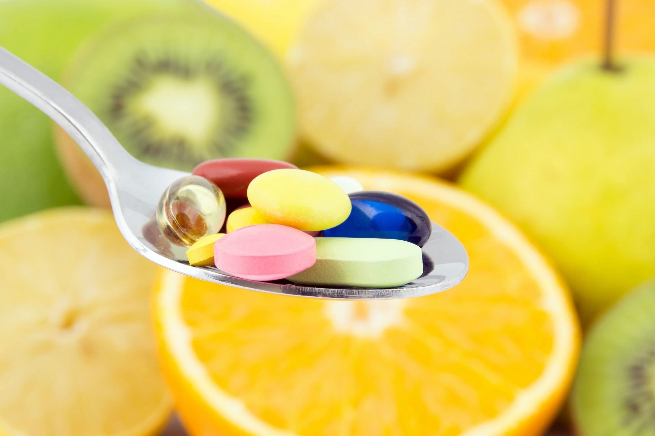 A spoonful of colorful vitamin pills with fresh fruit in the background.